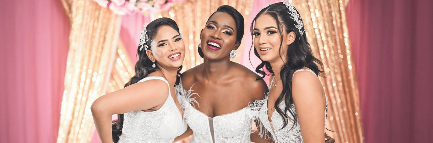 Three friends in white dresses laugh and embrace while posing together at a formal event with pink curtains behind them.