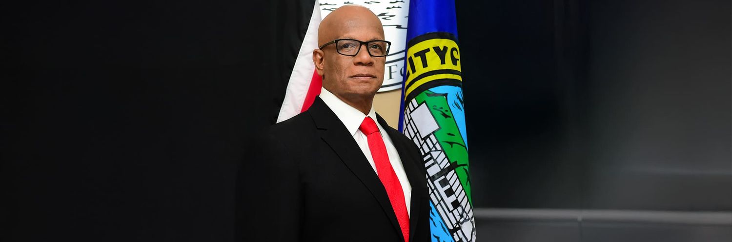 Person wearing glasses and red tie stands in front of an American flag and official government seal.