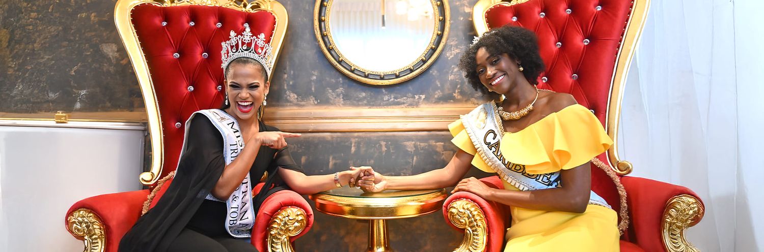 Two people laugh while sitting in ornate red velvet chairs with a large clock on the wall behind them.