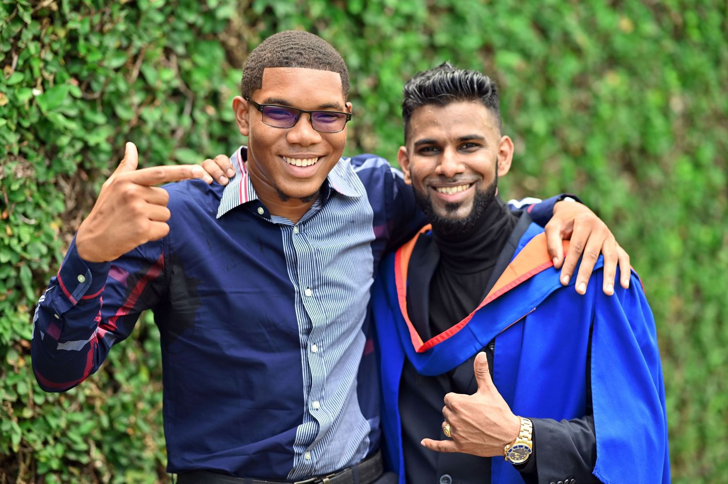 Two friends wearing casual attire giving thumbs up and smiling together outdoors with green foliage background.