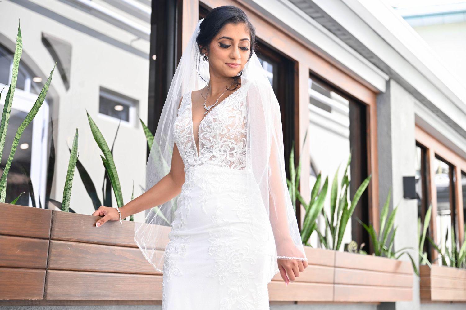 Bride in elegant white lace wedding dress with veil poses on outdoor terrace with modern architecture.