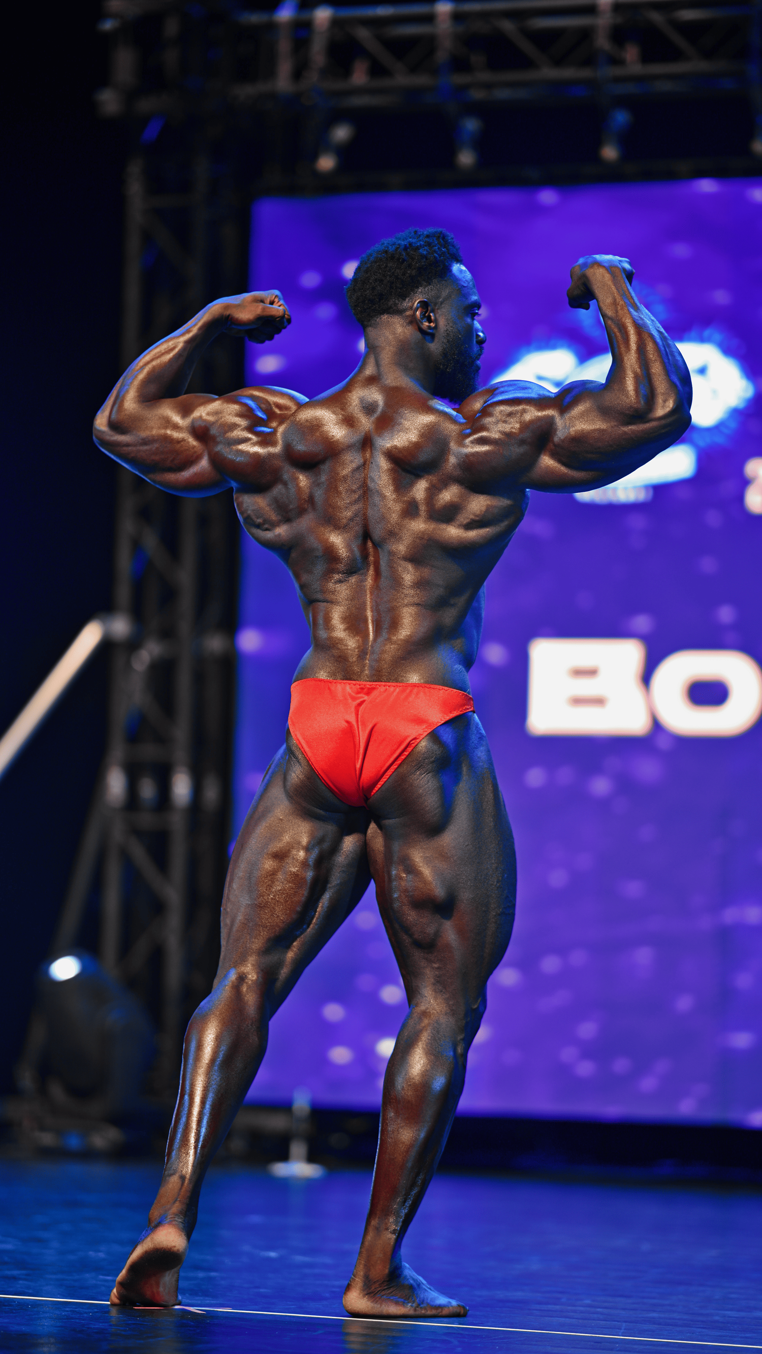 Bodybuilder performing rear double biceps pose showing defined back muscles on competition stage.