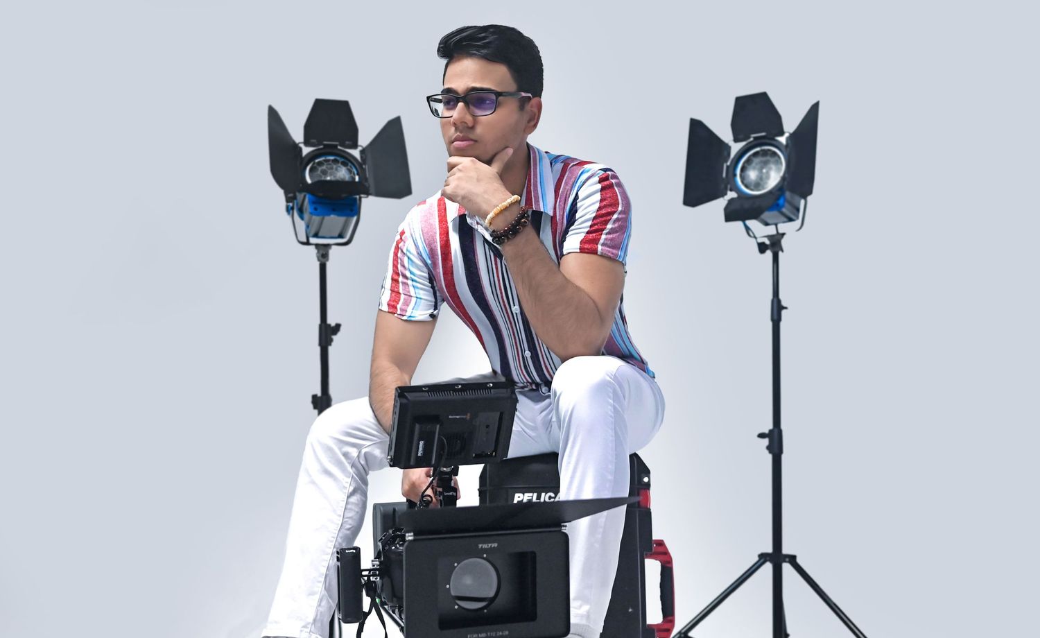 Professional videographer sitting with camera equipment and studio lights on white background.
