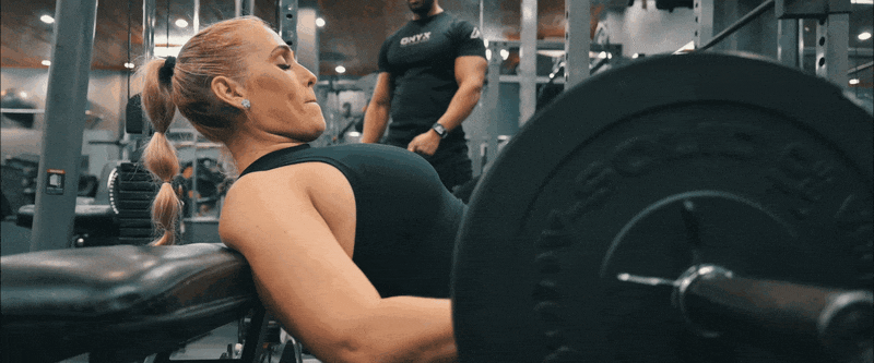 Woman performs hip thrust exercise with heavy barbell at a gym while positioned on weight bench.