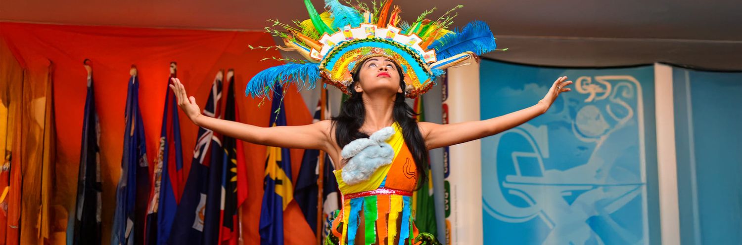 A performer in a vibrant Brazilian carnival-style costume with a feathered headdress dances with arms outstretched.