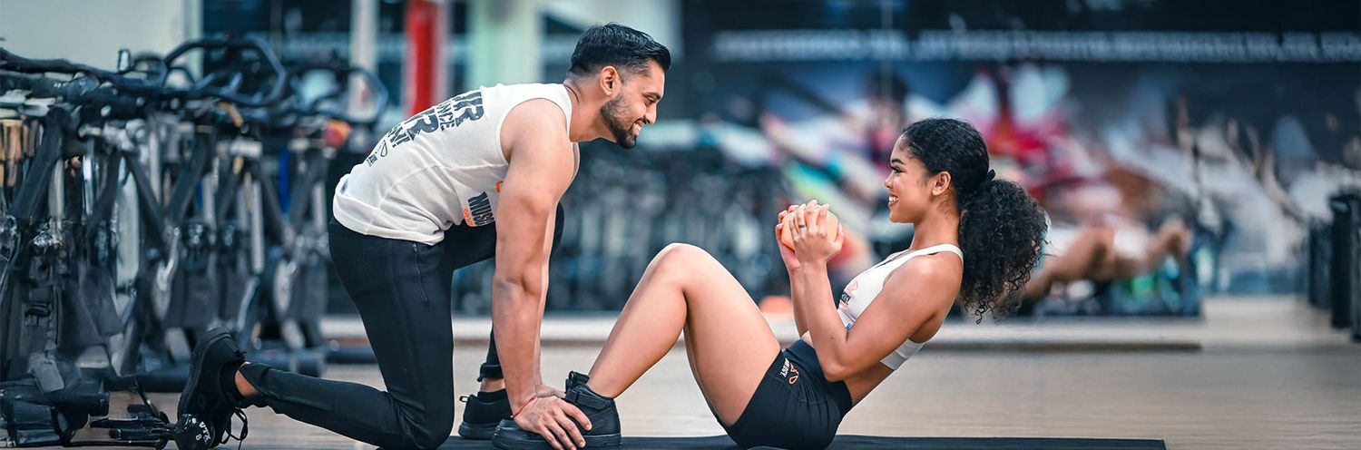 Two people exercising together in a gym with one assisting the other doing crunches on the floor.