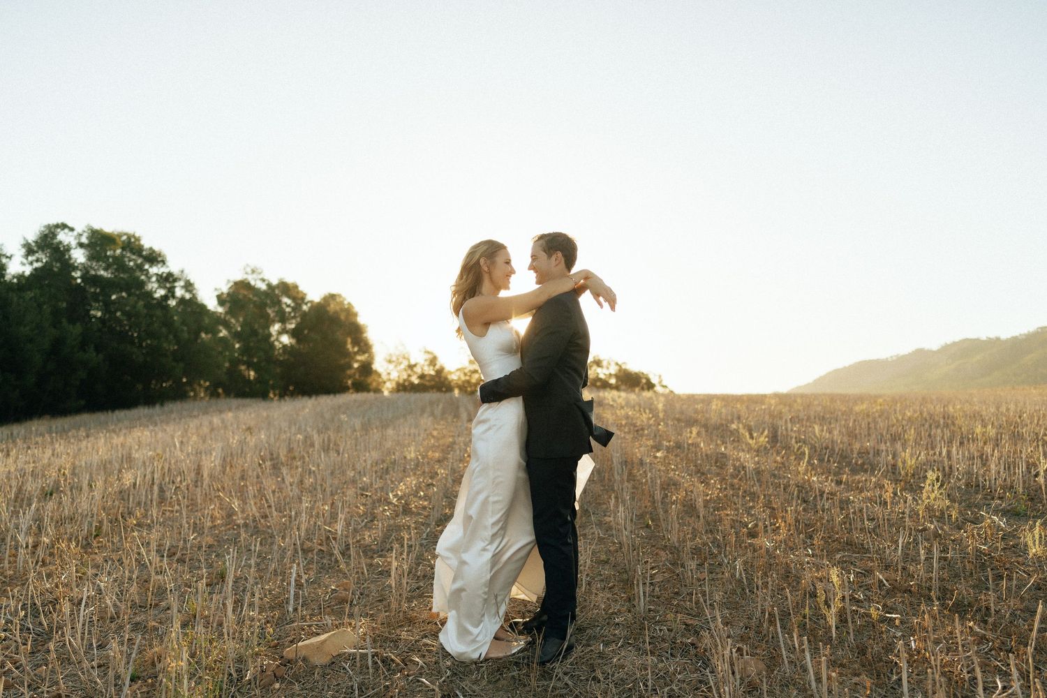 Bride and groom embrace in golden field at sunset, sharing a romantic dance during outdoor wedding photography session.
