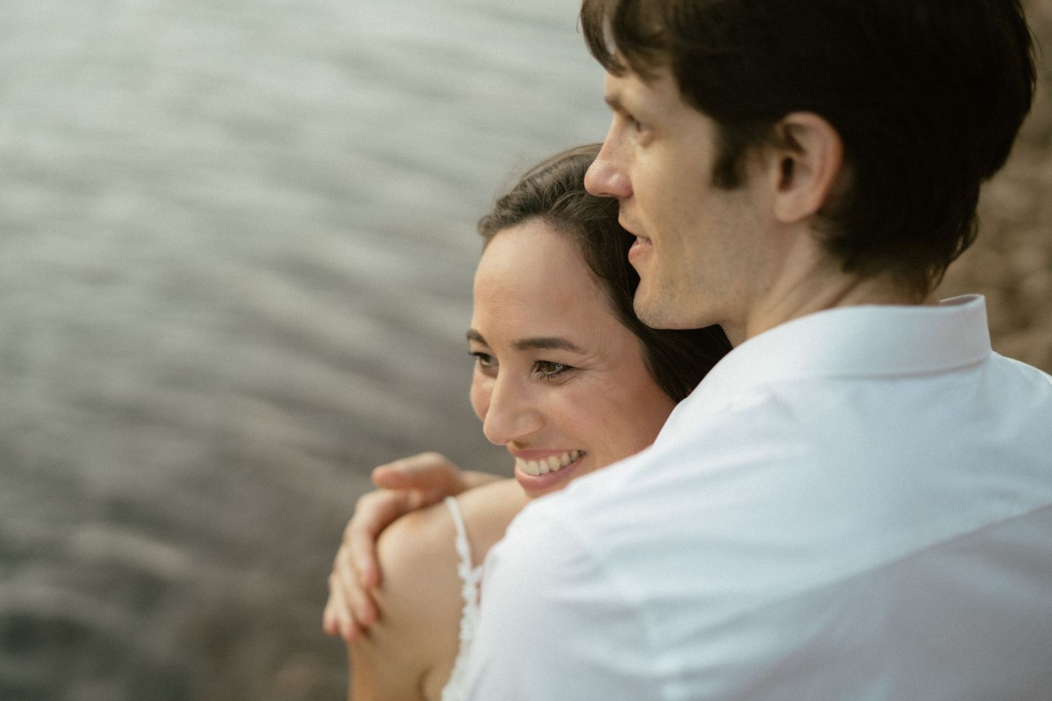 A smiling couple embraces near a serene waterfront, the woman in white laughing joyfully as the man holds her close.