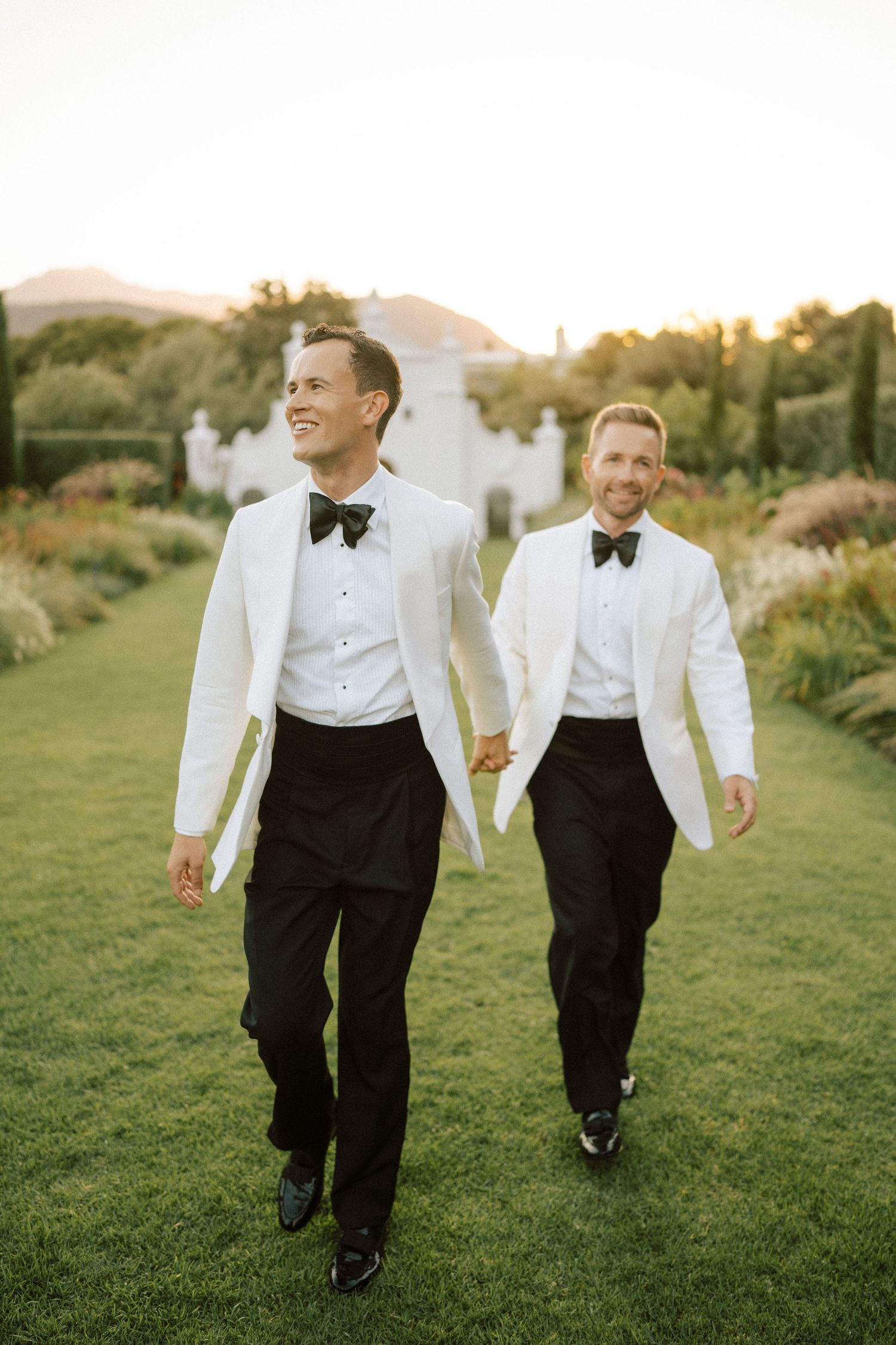 Two grooms in matching white tuxedo jackets and black bow ties hold hands walking across a green lawn at golden hour.