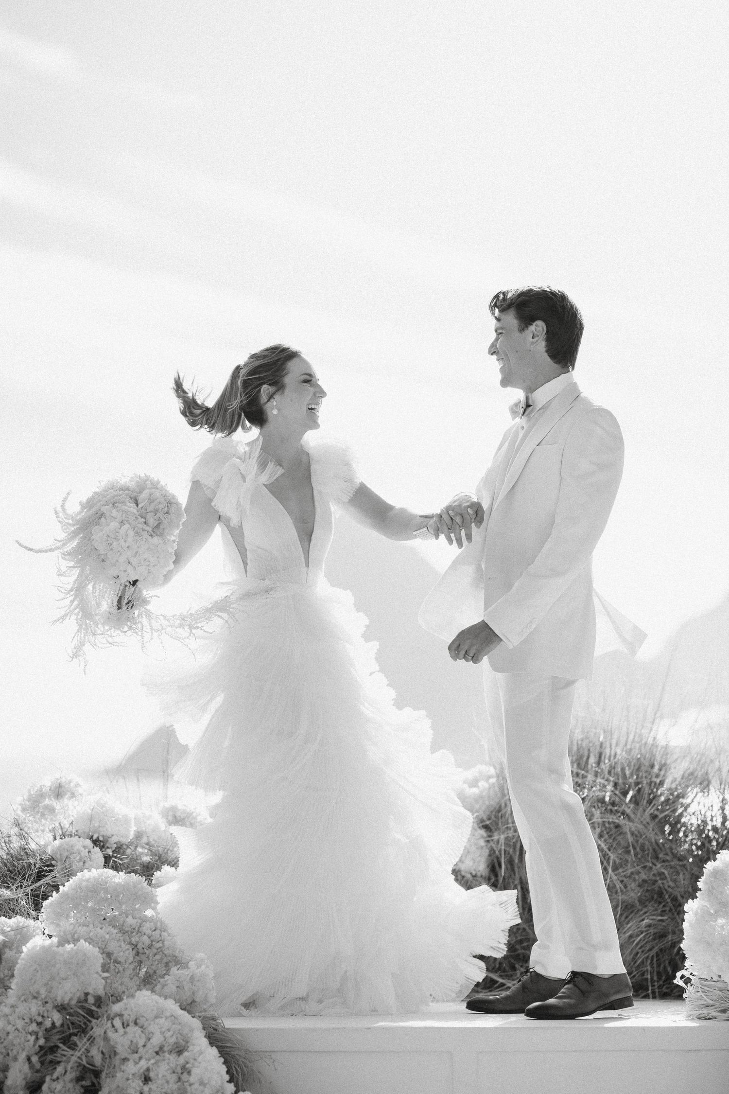 Black and white photo of a joyful bride and groom dancing outdoors, bride holding bouquet in flowing ruffled gown.