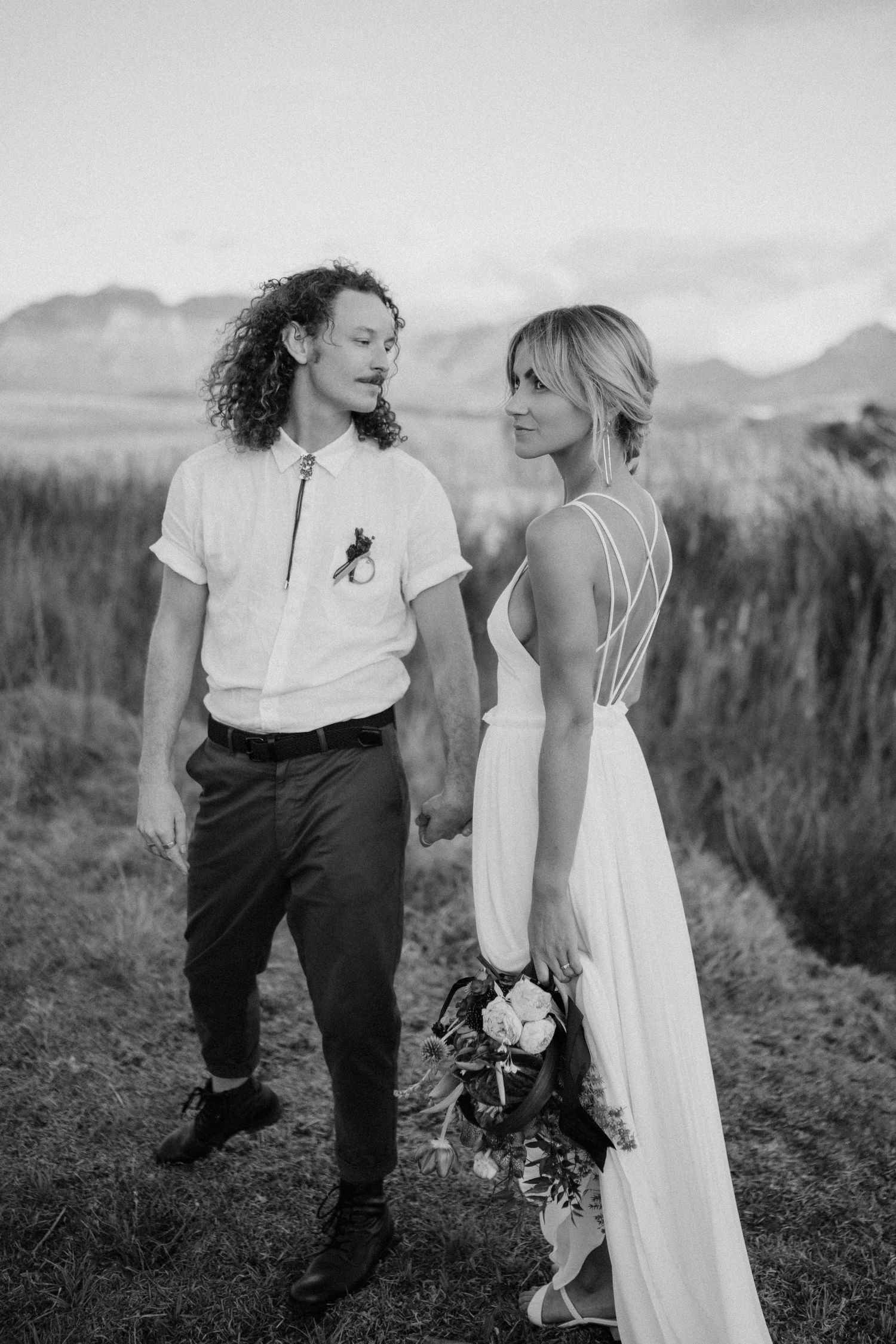 Black and white photo of a couple in wedding attire standing in a field with mountains in the background.