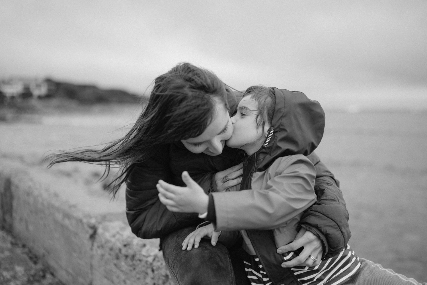 Black and white photo of a mother and young child sharing a tender kiss by the seaside on a cloudy day.