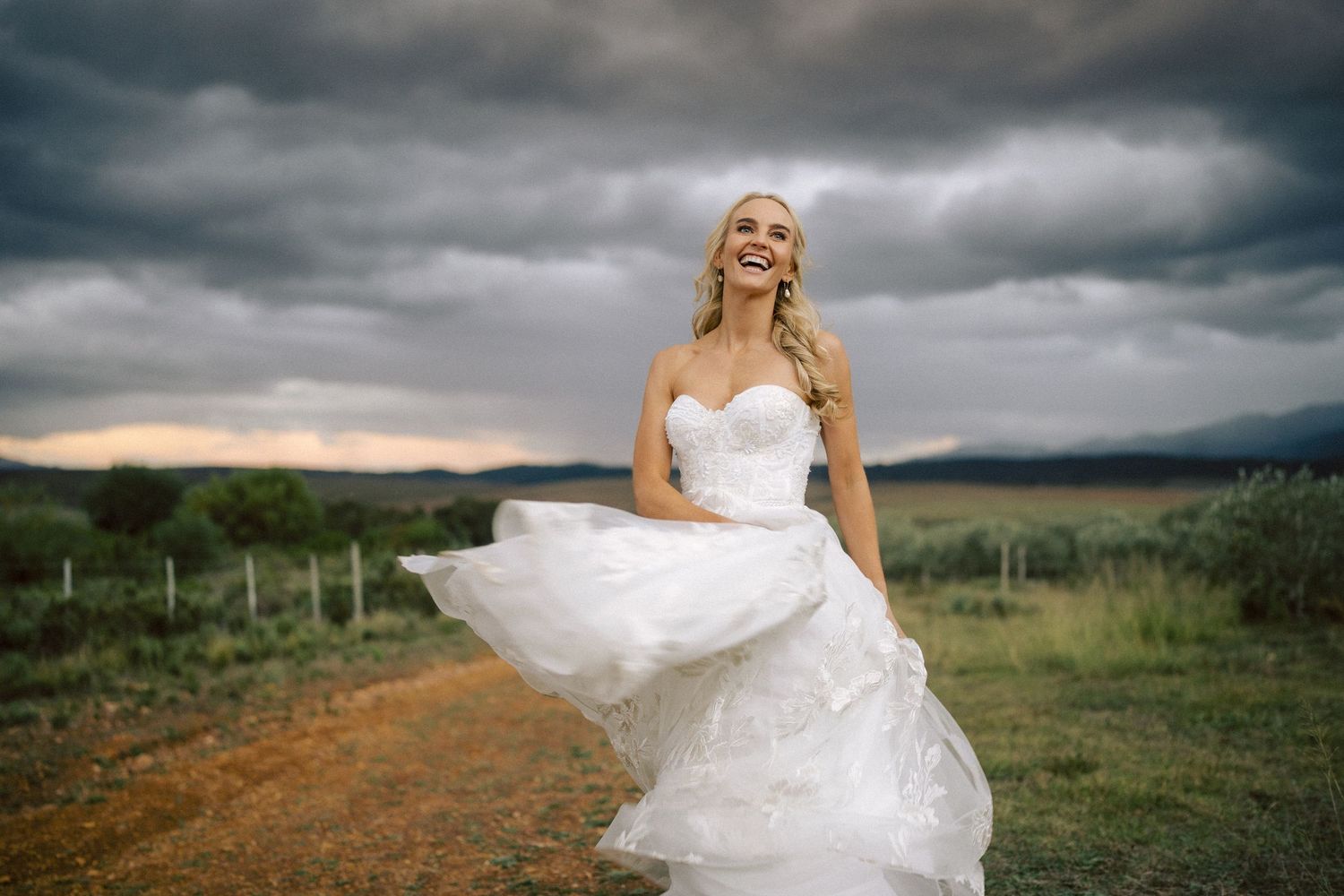 Bride in flowing white wedding gown laughing outdoors on a dirt path under dramatic cloudy skies.
