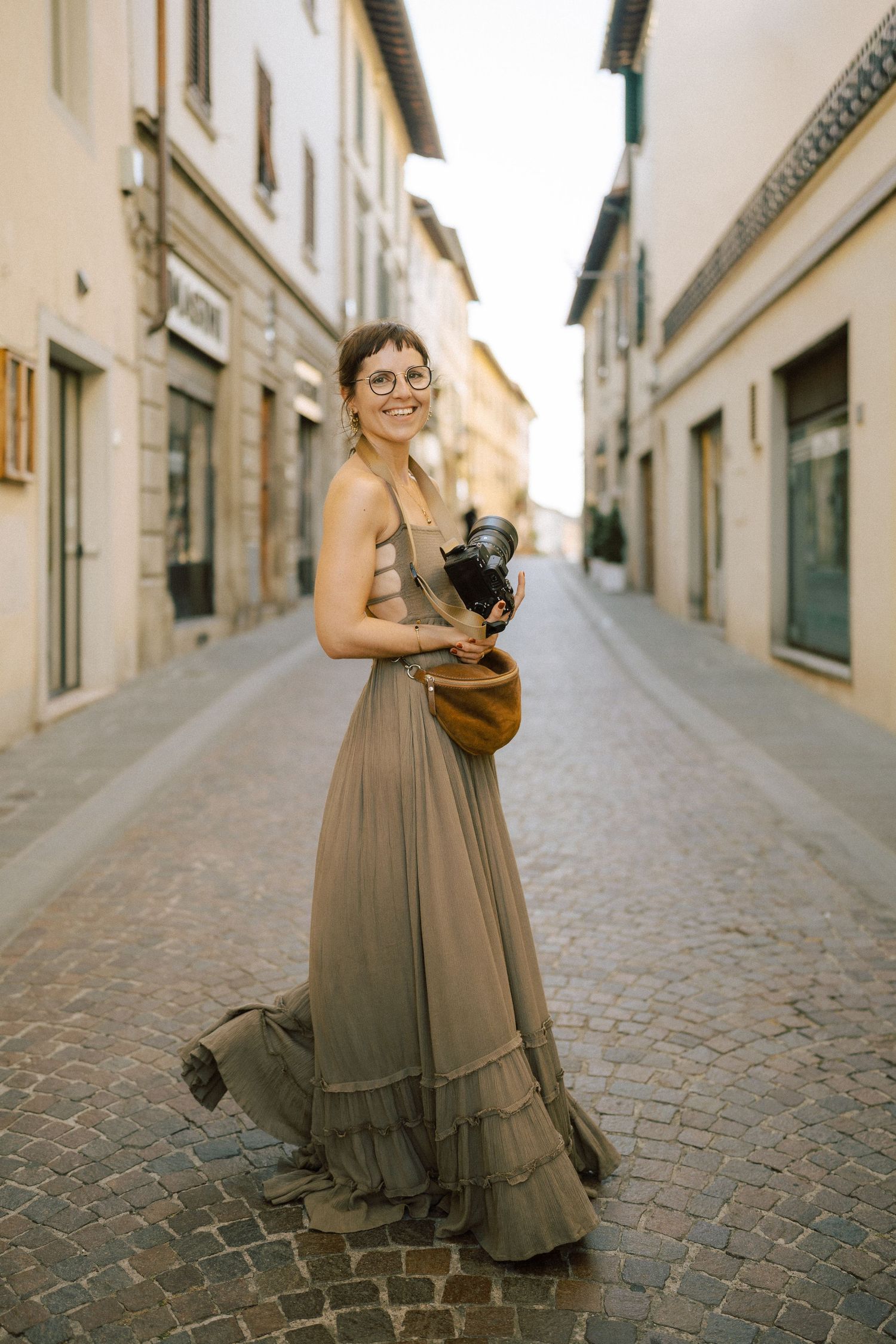 Smiling woman in olive maxi dress holding camera on cobblestone European street.
