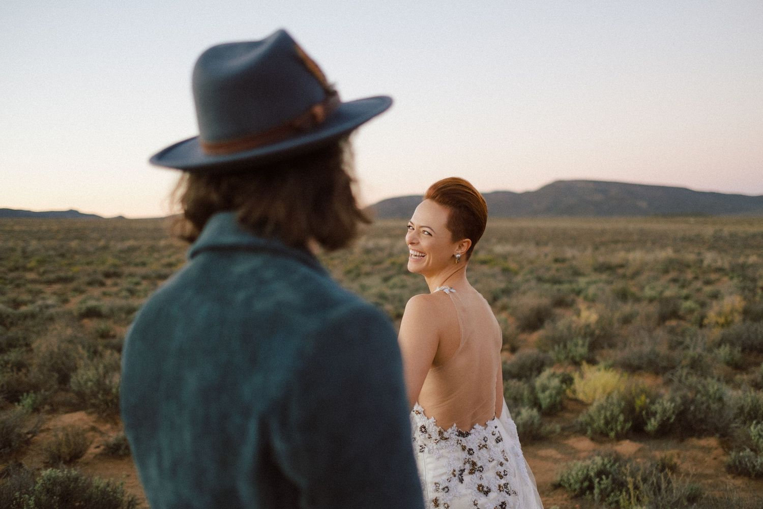 A smiling bride in a backless floral dress turns toward a man in a hat during a desert sunset engagement photo.