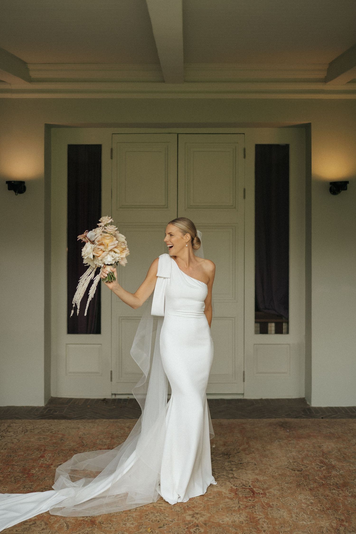 Bride in white one-shoulder gown holding bouquet, standing before elegant doors with wall sconces.