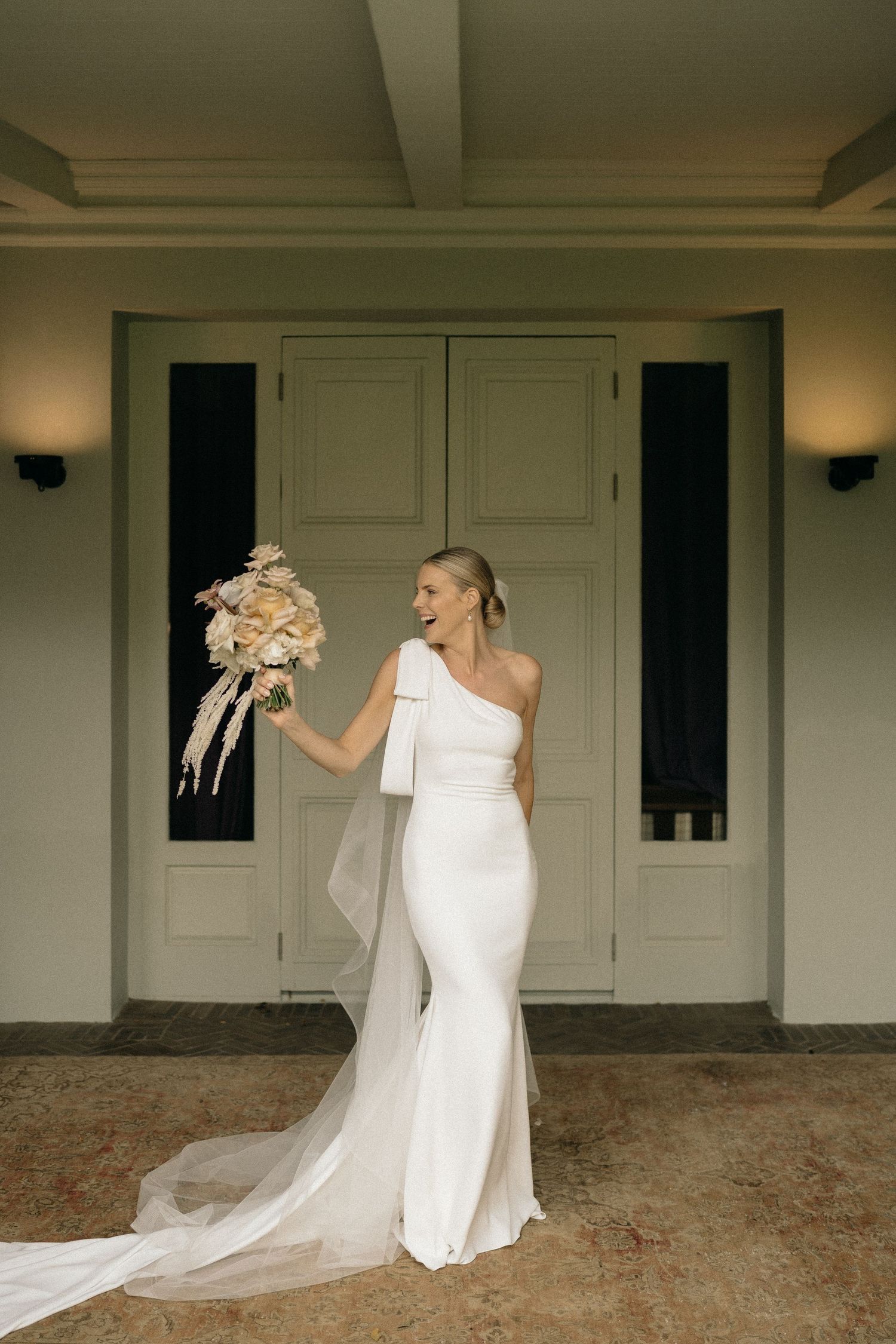 A bride in a white one-shoulder gown twirls playfully with her flowing veil against a neutral building exterior.