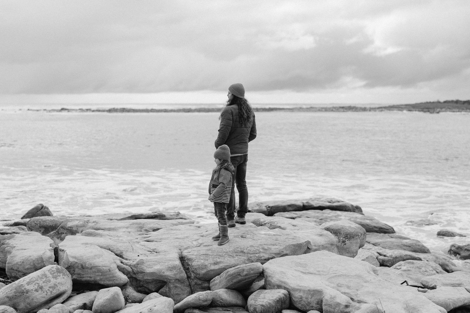 Black and white photo of a parent and child standing on rocky shore, gazing at the ocean on a cloudy day.