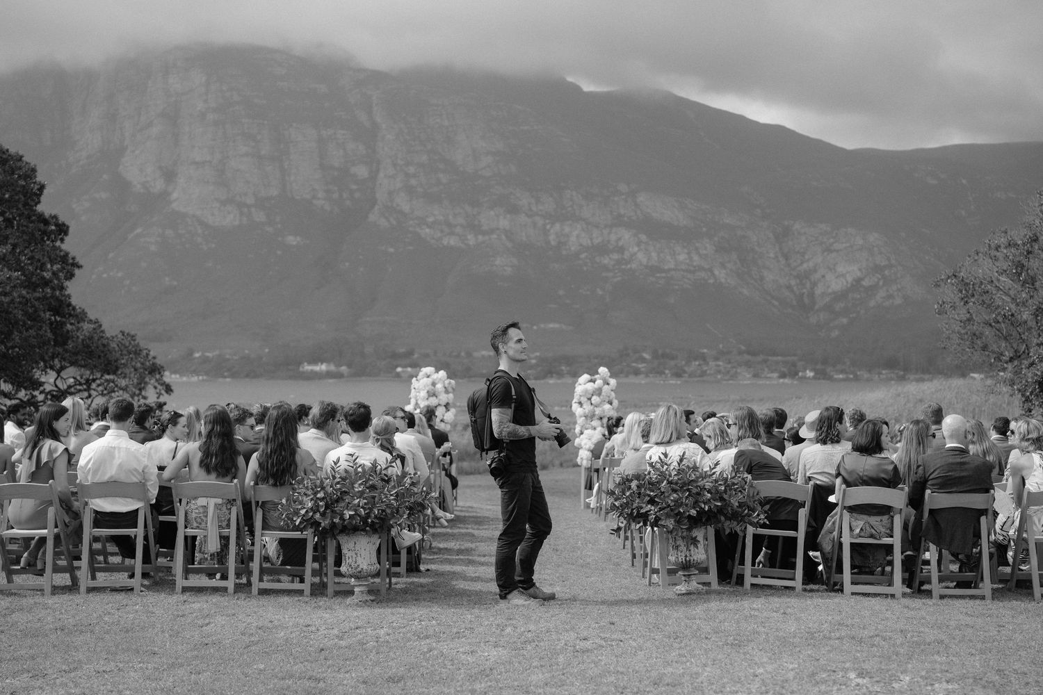 Black and white outdoor wedding ceremony with guests seated facing an officiant, mountains and lake in the background.