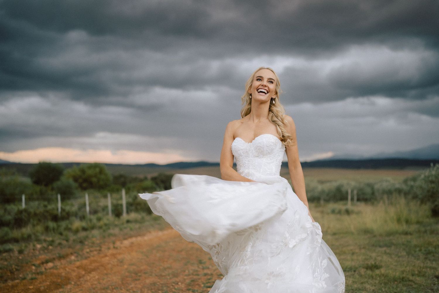 Smiling bride in flowing white lace wedding dress twirling outdoors under dramatic stormy sky in rural landscape.