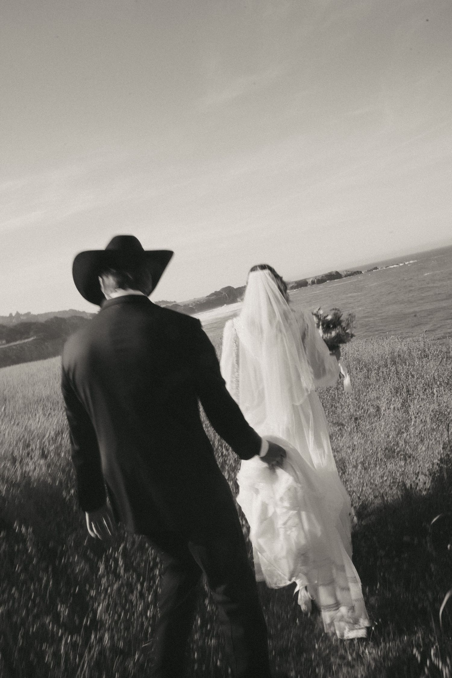 Groom walks with bride, holding her vintage thrifted wedding dress as they stroll through a field in Sea Ranch, CA