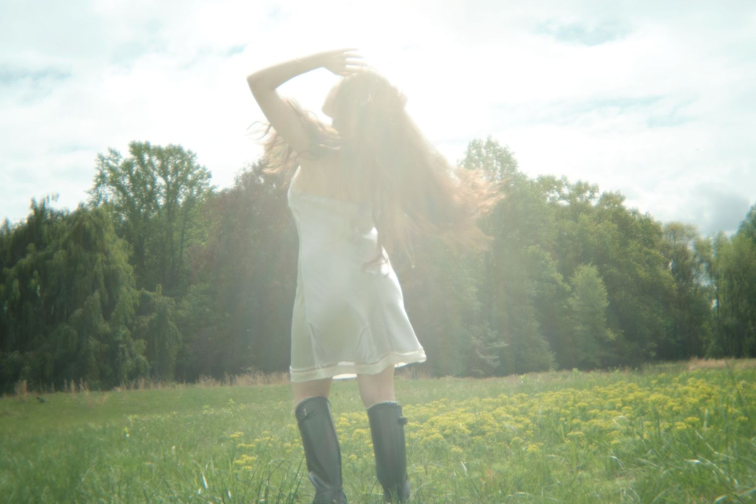 Figure in a white dress and boots poses in a sunlit meadow with trees in the background.
