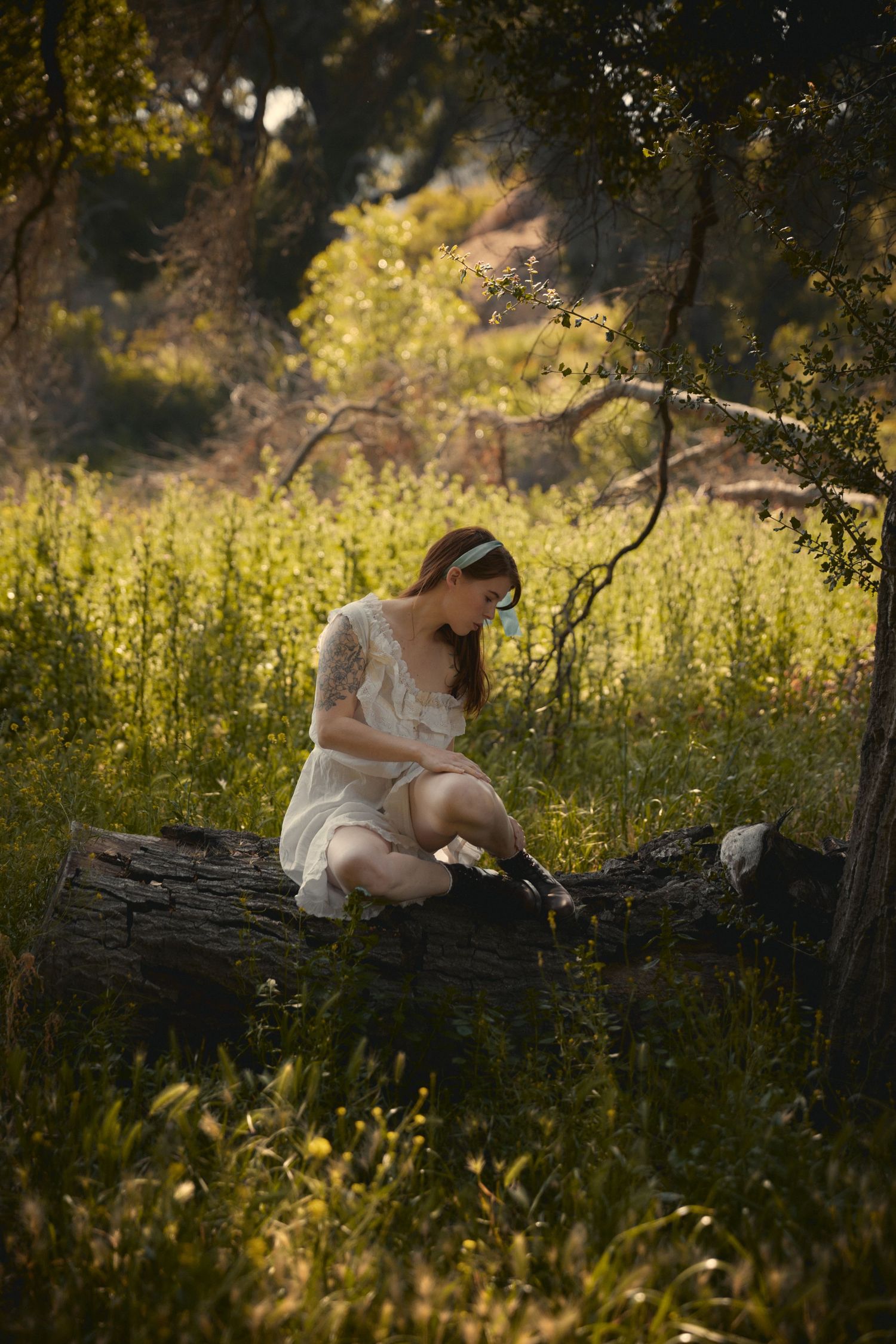 Dreamy sunlit scene of a woman in white dress sitting on fallen log among wildflowers.