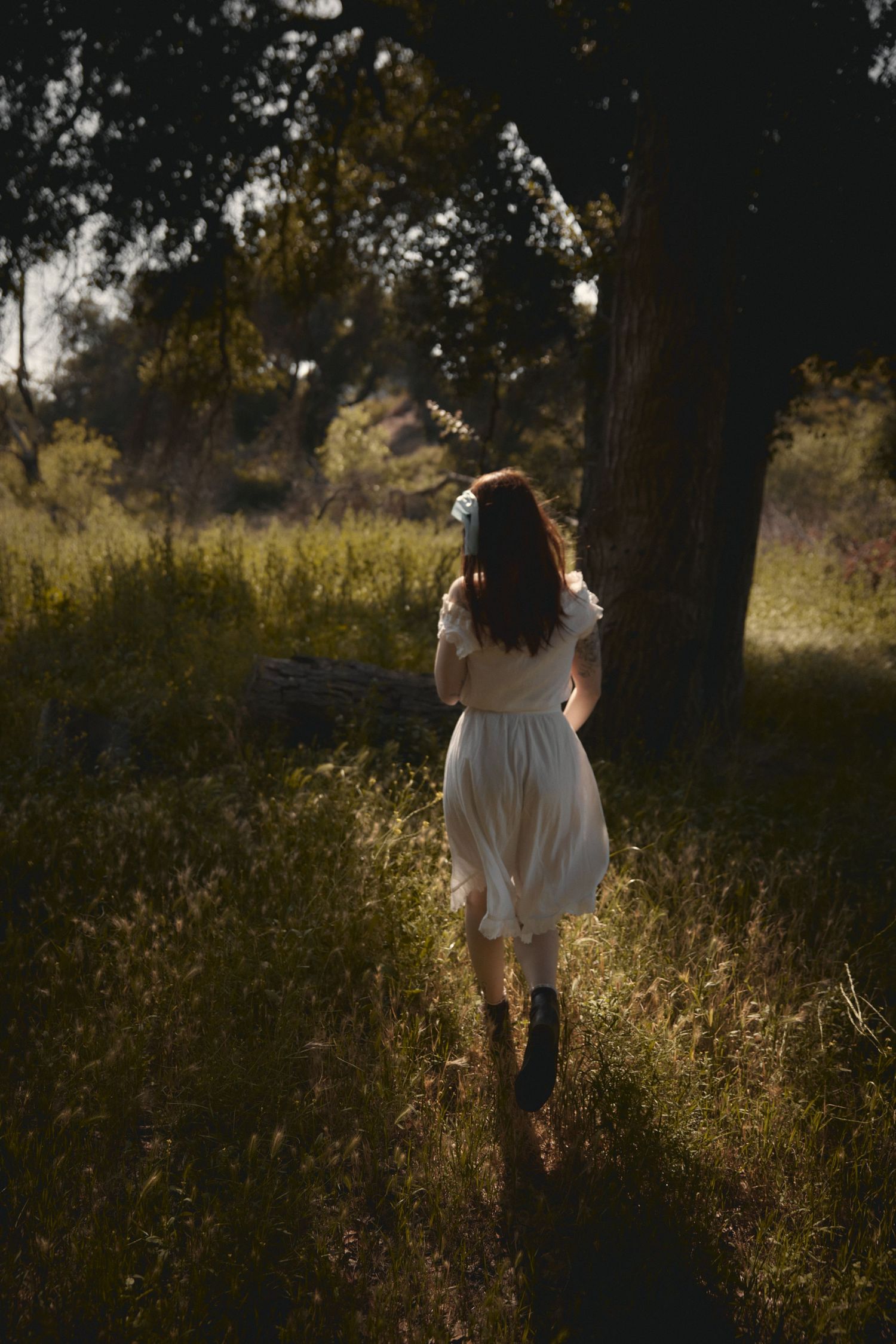 Dreamy backlit figure in white dress walking through tall grass at golden hour.
