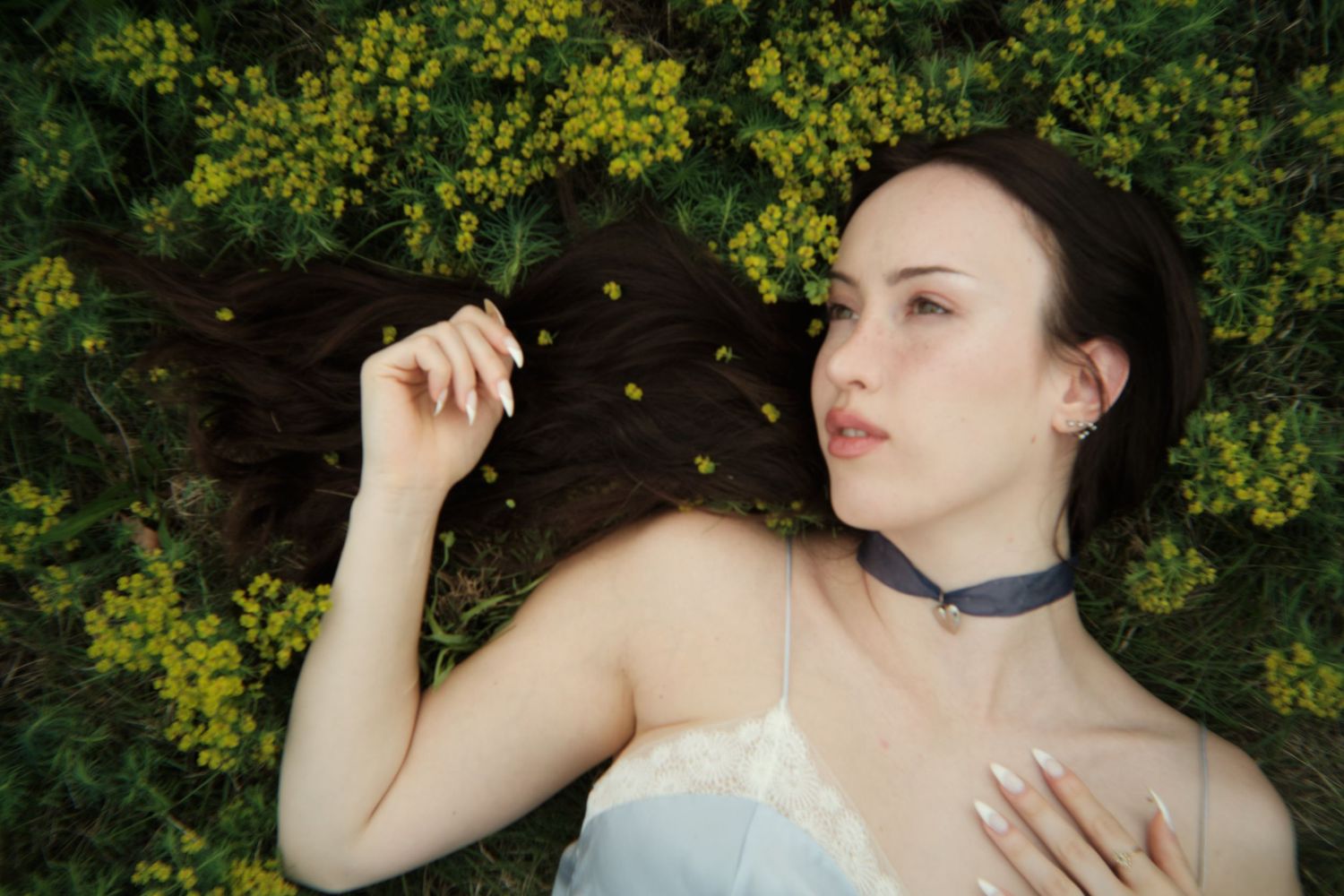 Figure in white dress lies among yellow flowers with dark hair spread on ground and blue choker necklace.