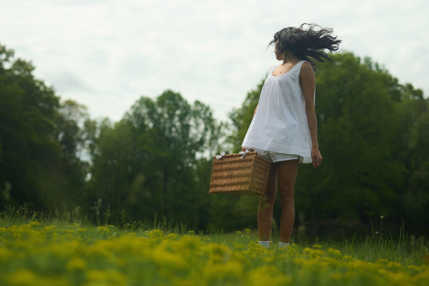 A person in a white dress stands in a field of yellow wildflowers holding a wicker basket on a summer day.