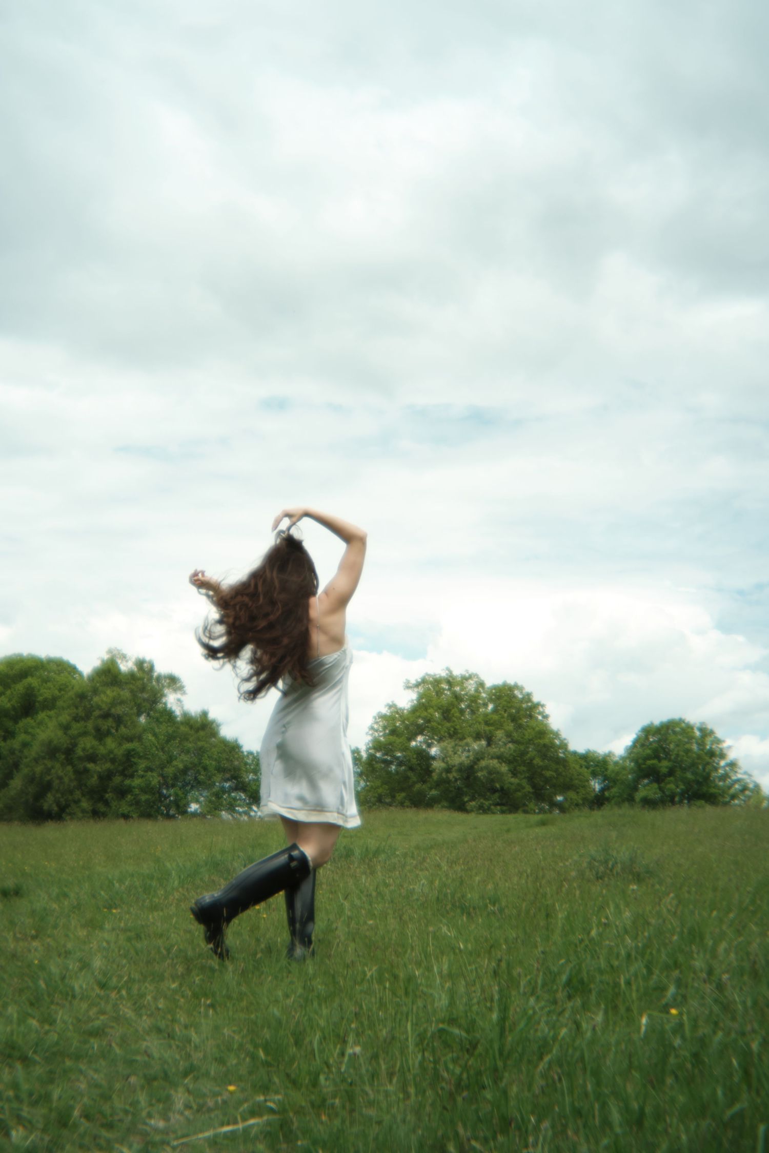 A dreamy figure in a white dress and dark boots dances in a green meadow under cloudy skies.