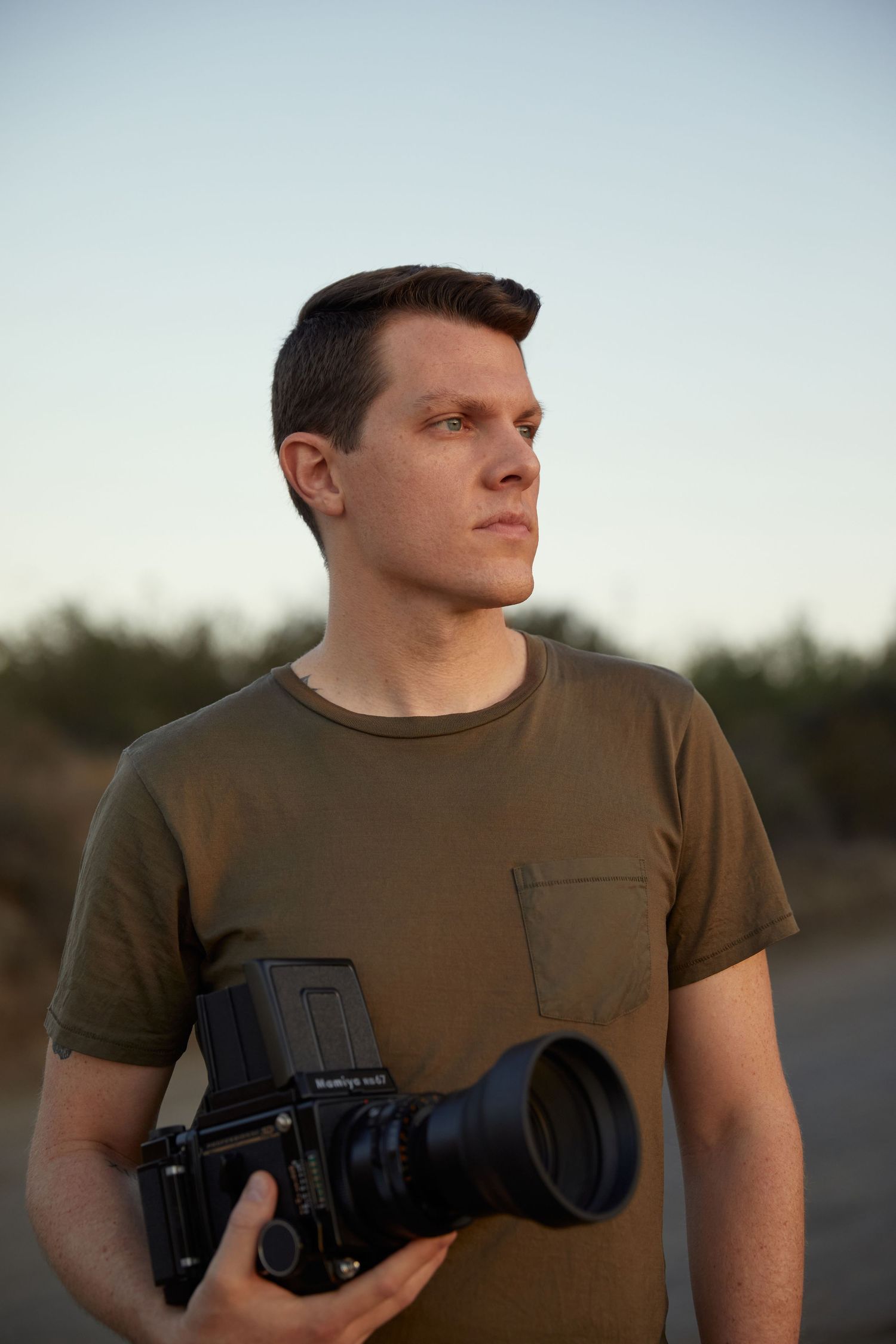 Adam Wamsley, photographer, wearing a green t-shirt holds a medium format film camera at dusk.