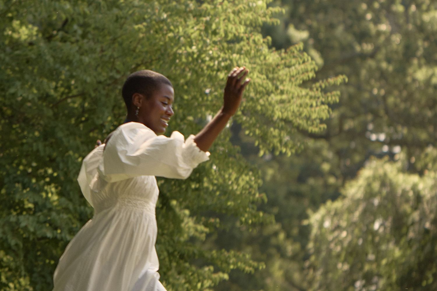 A figure in a flowing white dress dances gracefully in a sunlit outdoor setting with trees in the background.