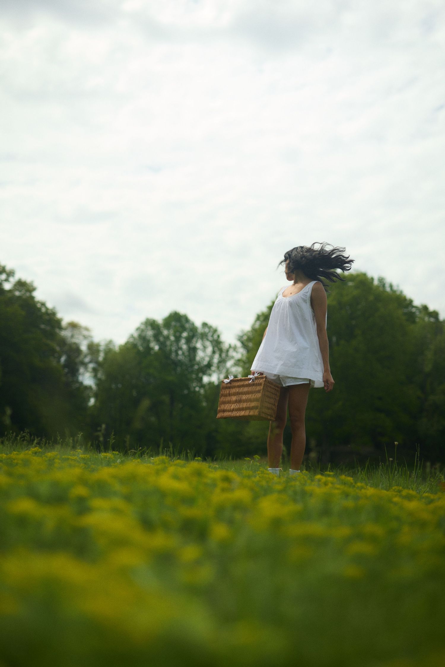 A person in a white dress stands in a field of yellow wildflowers holding a wicker basket on a cloudy day.