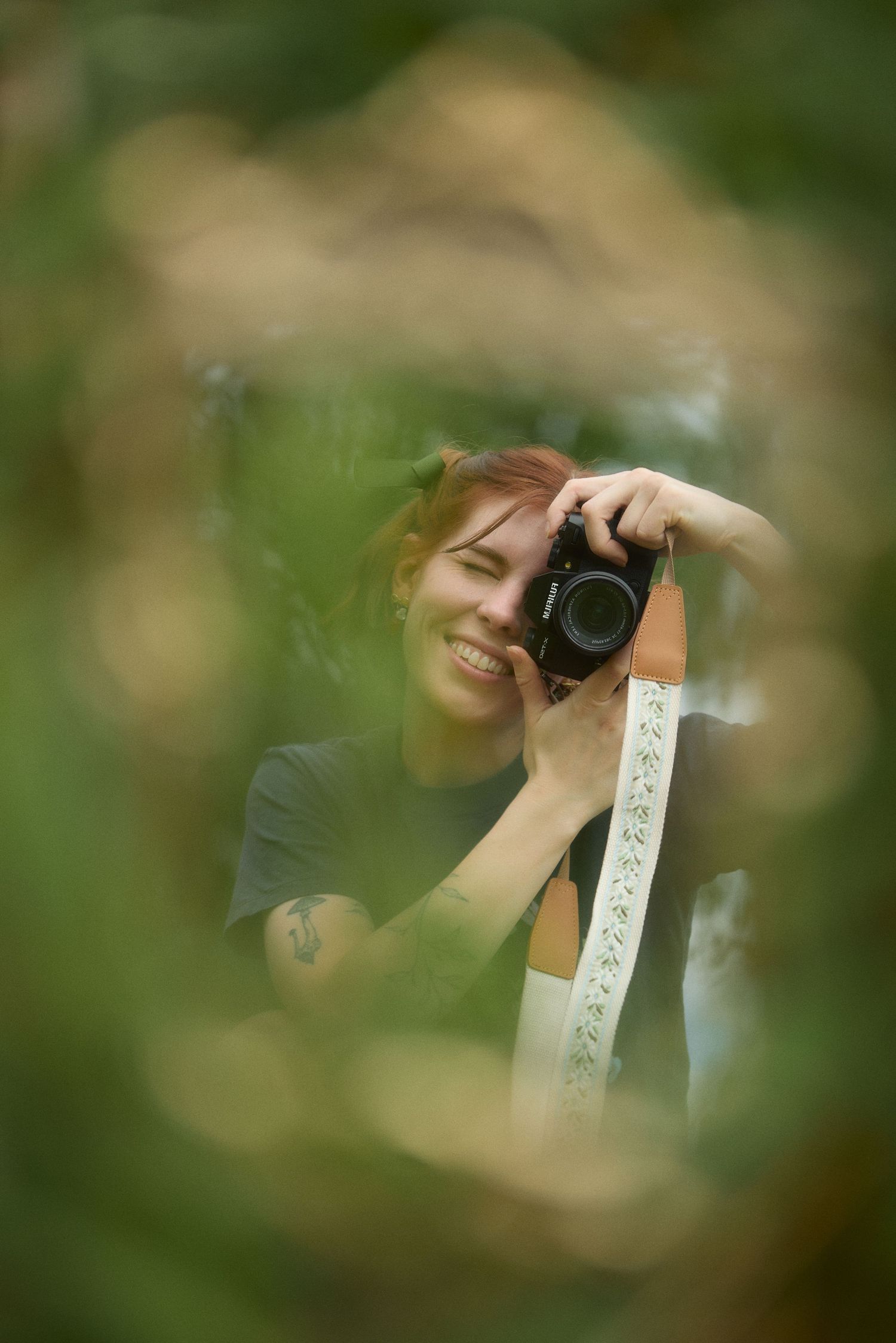 Through blurred green foliage, a photographer wearing dark clothing holds up a camera to take a picture.
