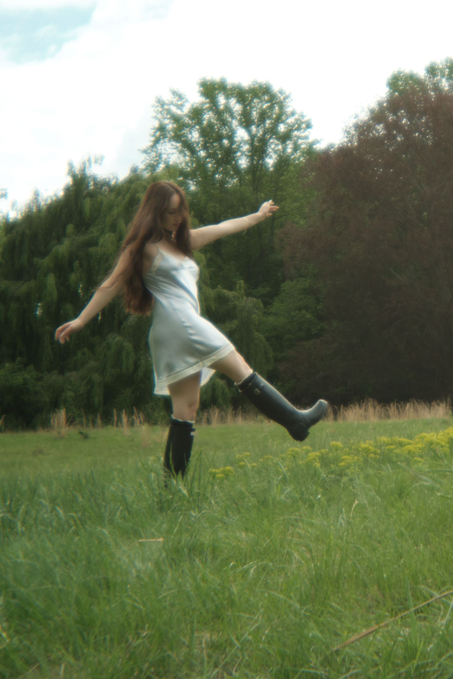 A figure in a white dress and rain boots strikes a playful pose while dancing in a grassy meadow.