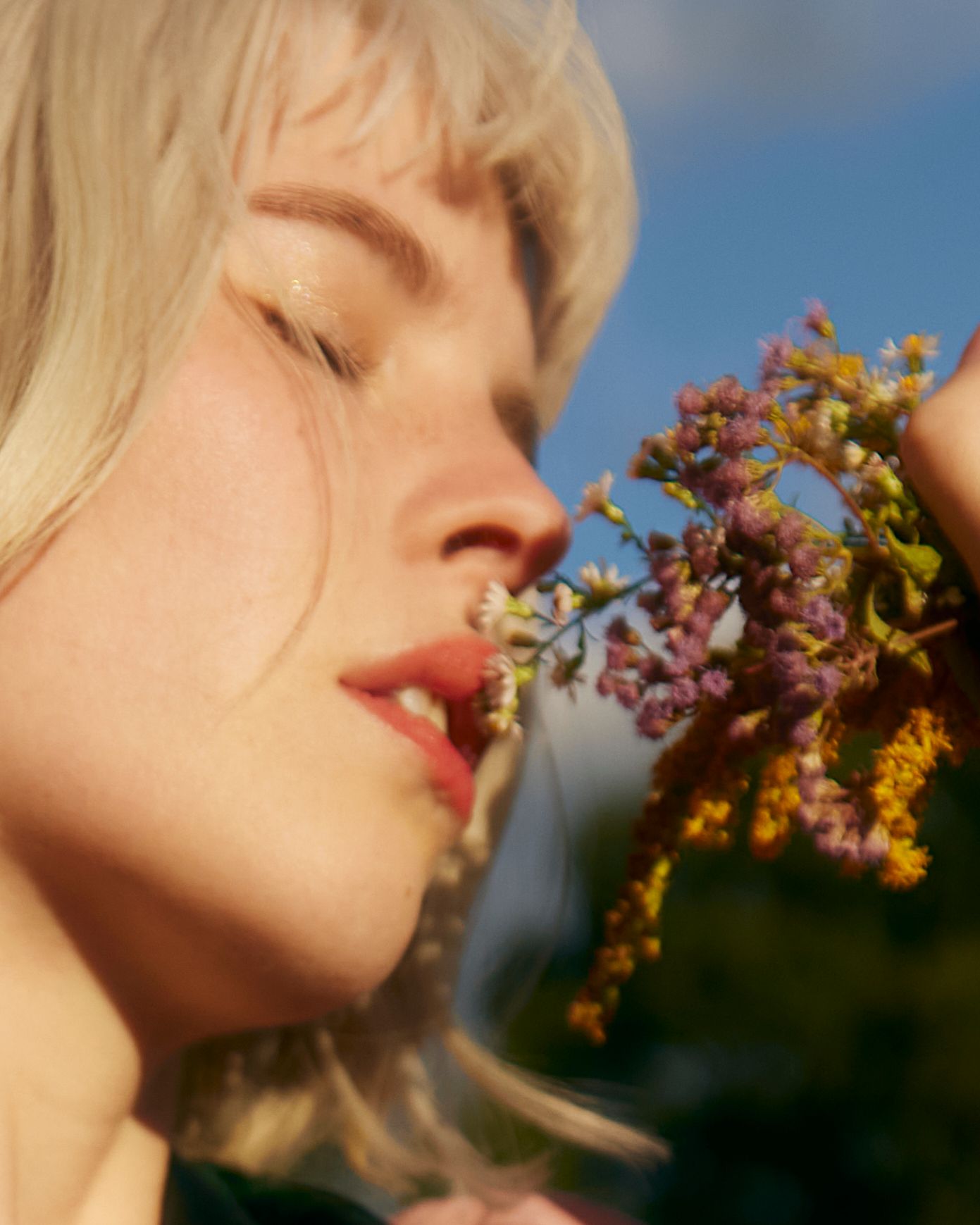 A close-up profile shot shows someone smelling delicate purple flowers in warm sunlight.