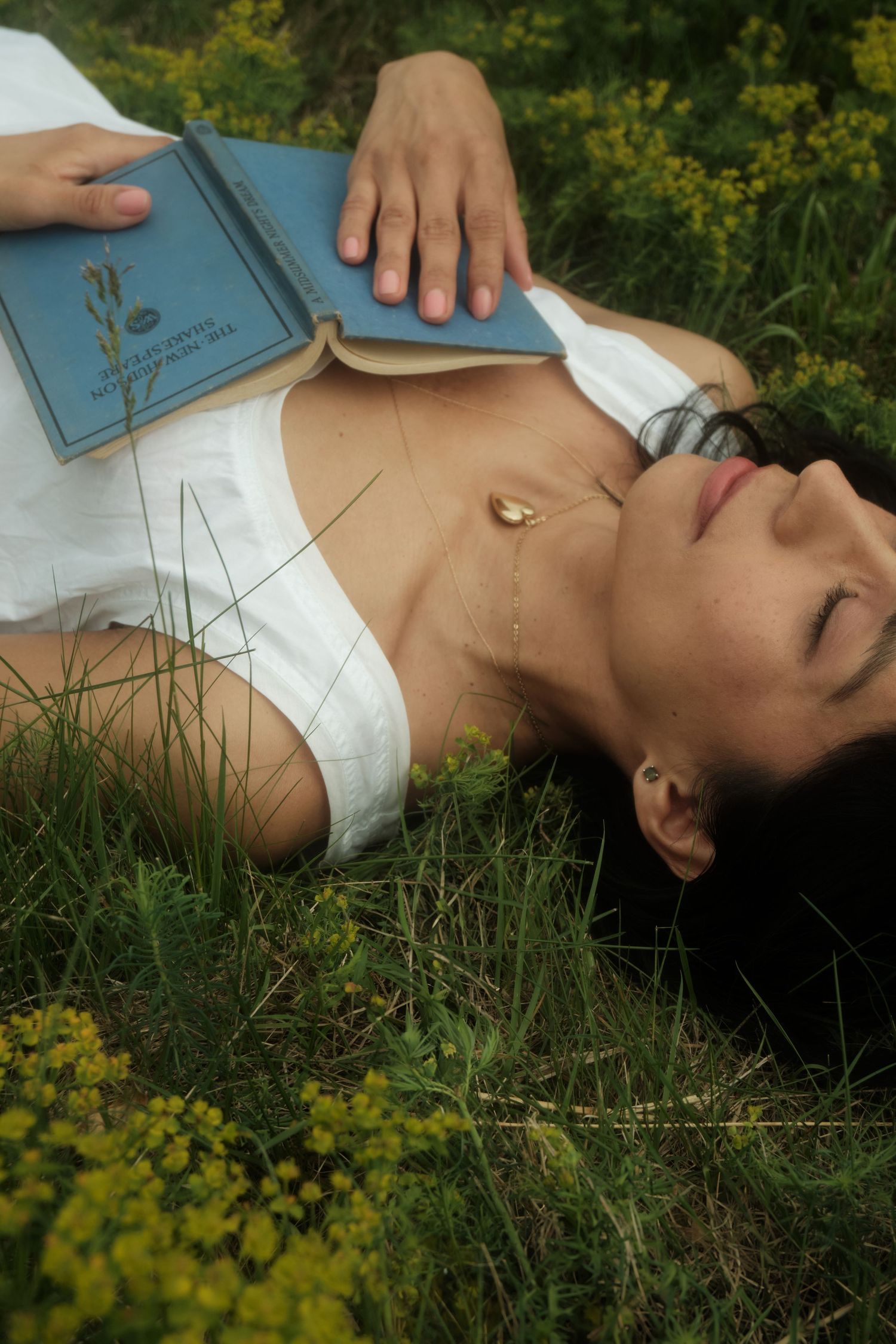 Someone lies peacefully in the grass among yellow wildflowers holding a blue book on a sunny day.