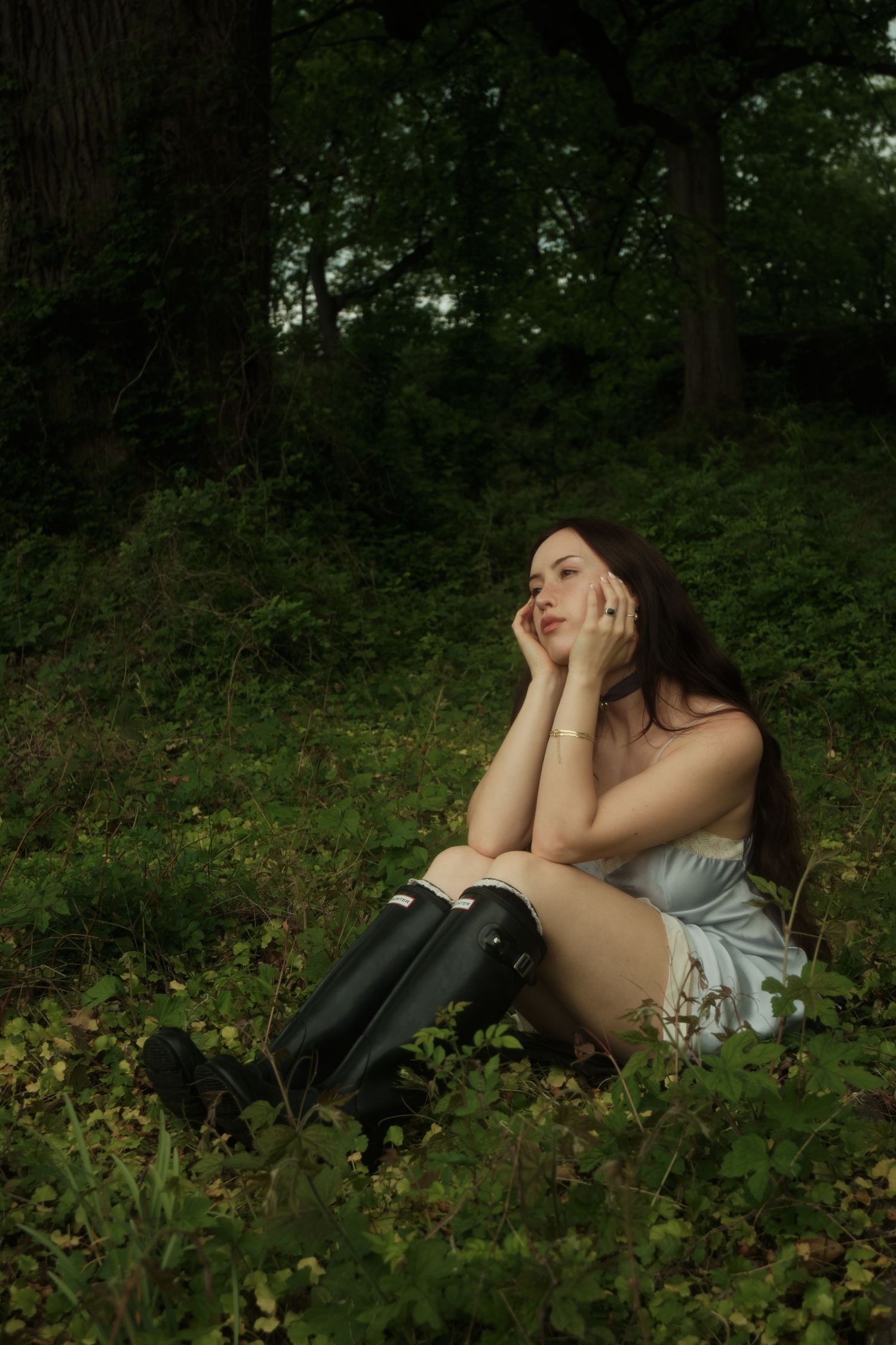 A dreamy forest scene shows someone in a light dress and tall boots sitting thoughtfully among green foliage.
