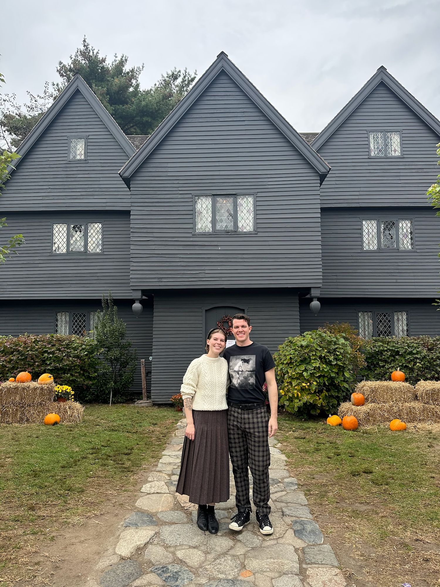 A couple poses in front of a dark gray historic house decorated with pumpkins for fall.