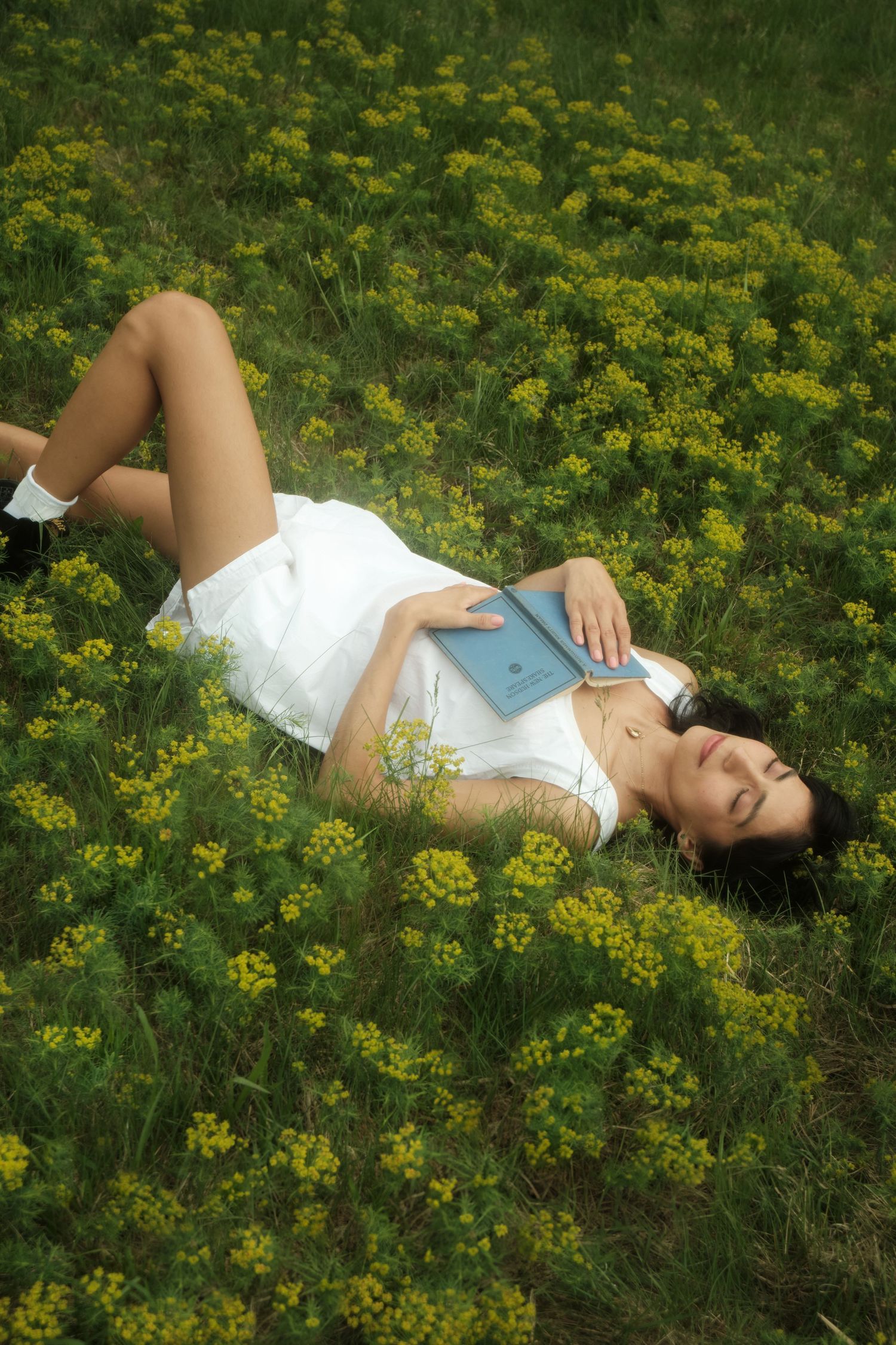 Person in white dress lying in field of yellow wildflowers with book on summer day.