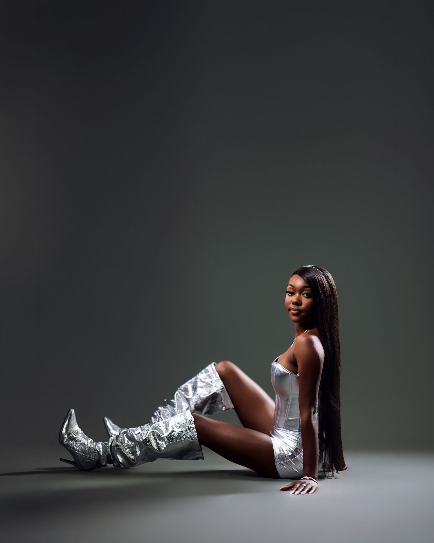 A woman in a silver metallic outfit and knee-high boots sits on a studio floor, posing confidently.