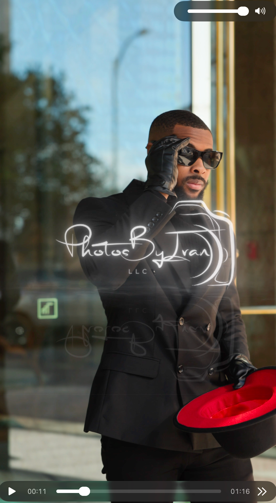 Man in a black suit and leather gloves adjusts dark sunglasses while holding a red-lined black fedora in front of a glass storefront.