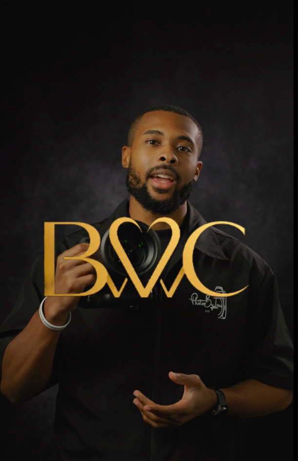 Bearded man in a black embroidered shirt gestures while holding a camera, with a golden BWC heart logo overlaid against a dark backdrop.