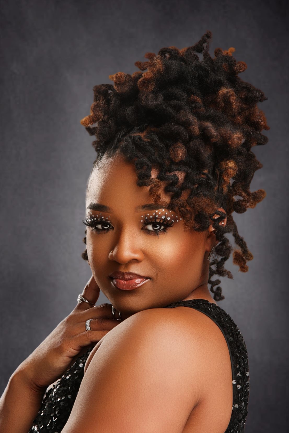 A Black woman with styled locs updo, decorative eye makeup, and a black sequined dress poses against a gray background.