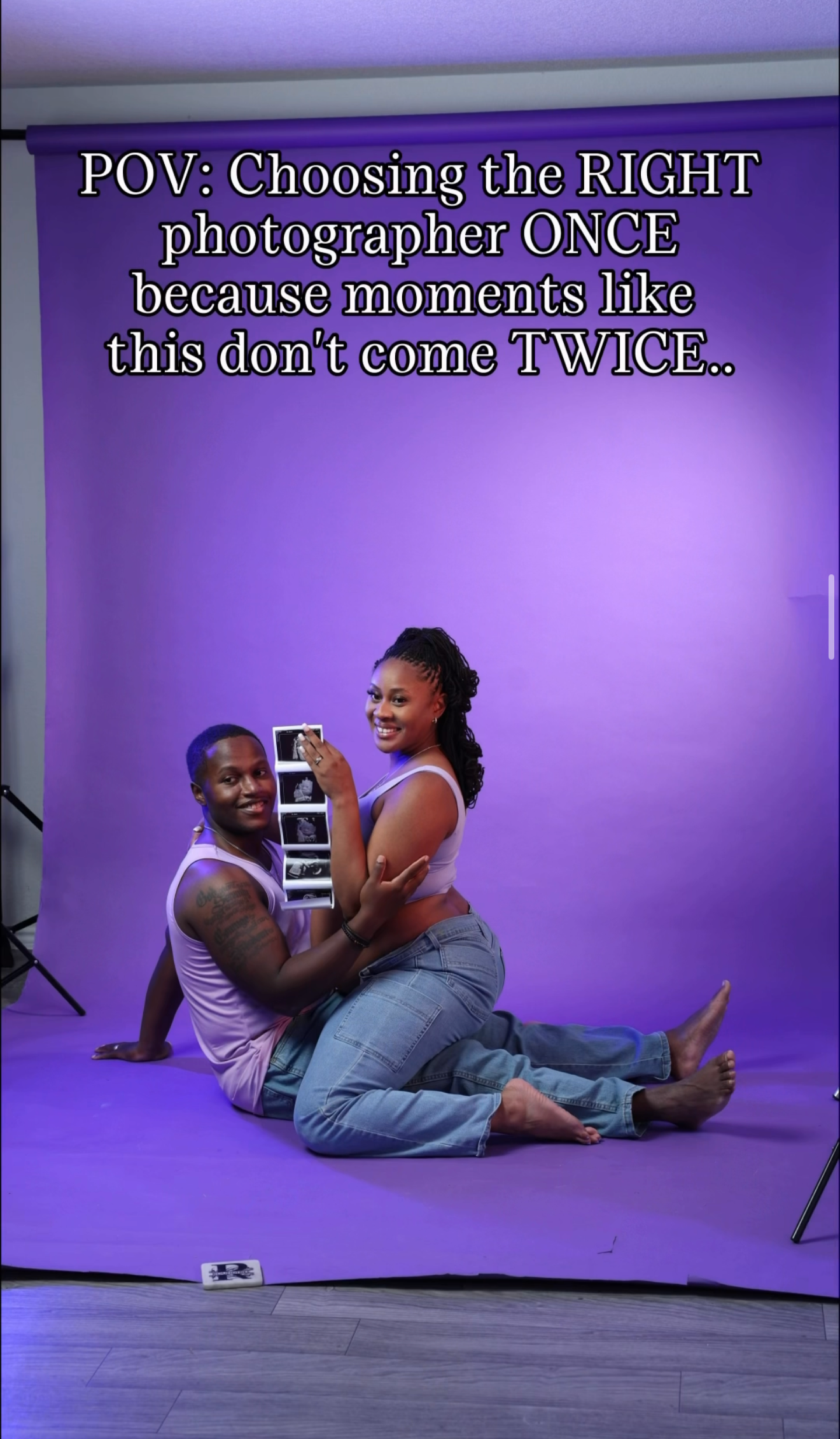 Smiling couple in jeans and tank tops sit together on a purple studio backdrop holding an ultrasound photo in a maternity photoshoot.