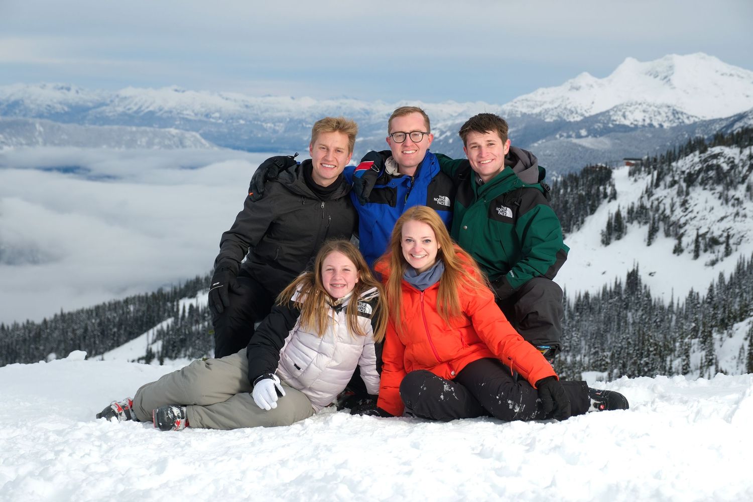 Five friends pose together on a snowy mountain slope with evergreen forest and cloud-filled valley below.