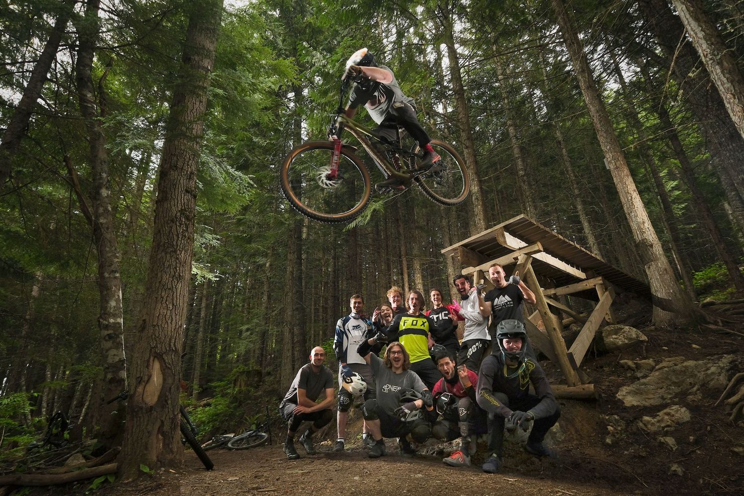 Mountain biker performs aerial jump over wooden ramp in forest while spectators watch from below.