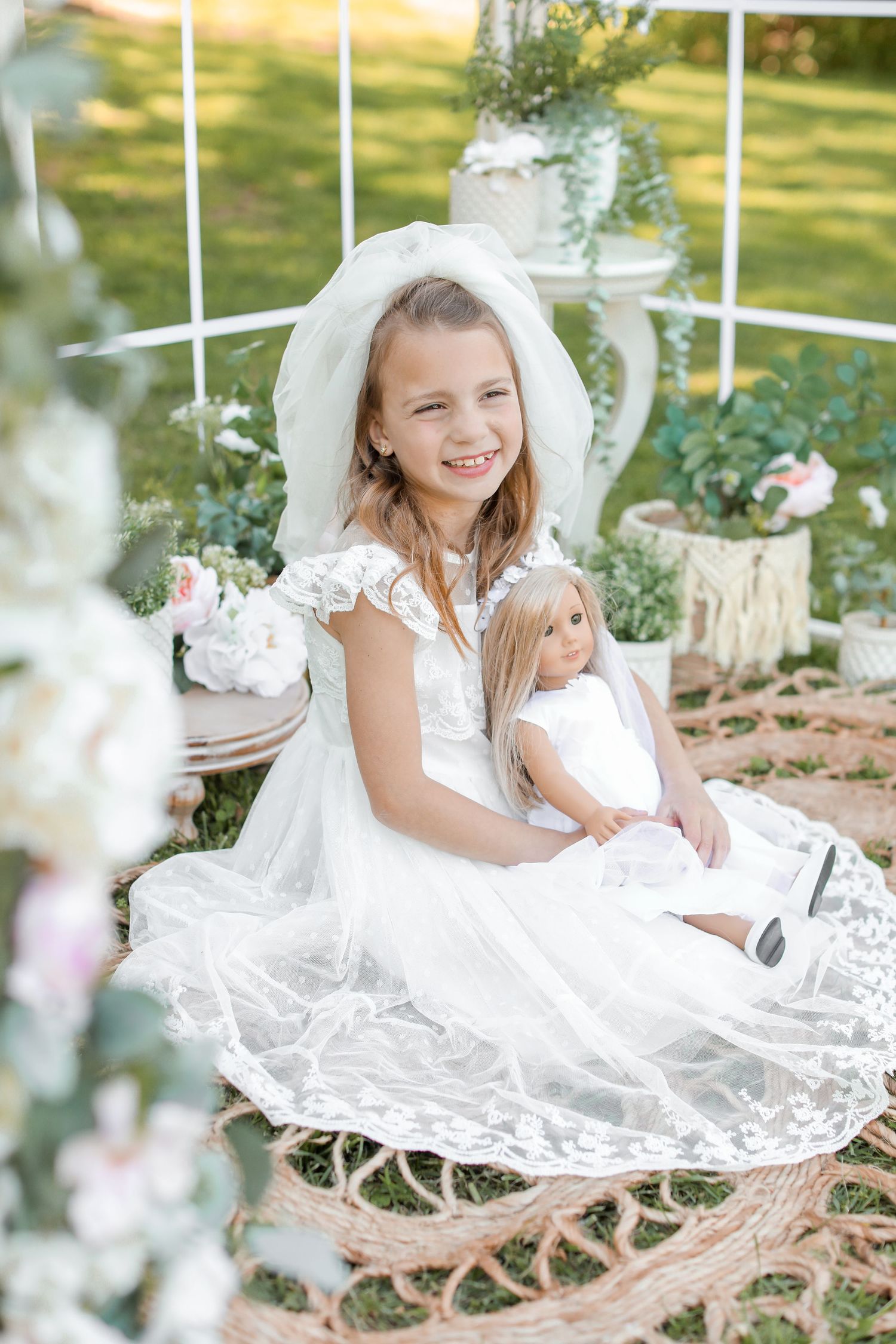 Child in white dress with doll sitting in garden swing surrounded by flowers.