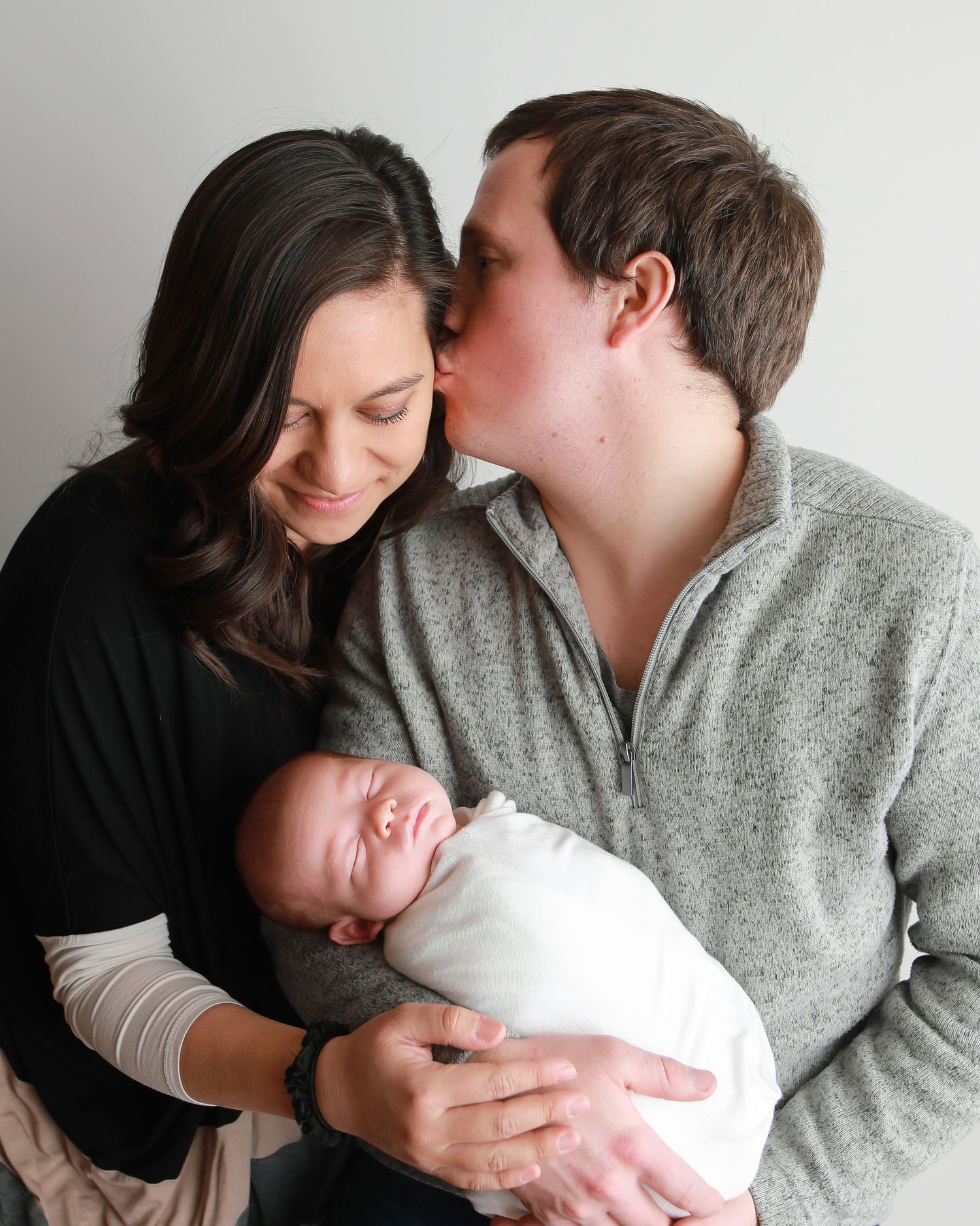 A couple shares an intimate moment while holding their sleeping newborn baby wrapped in white.
